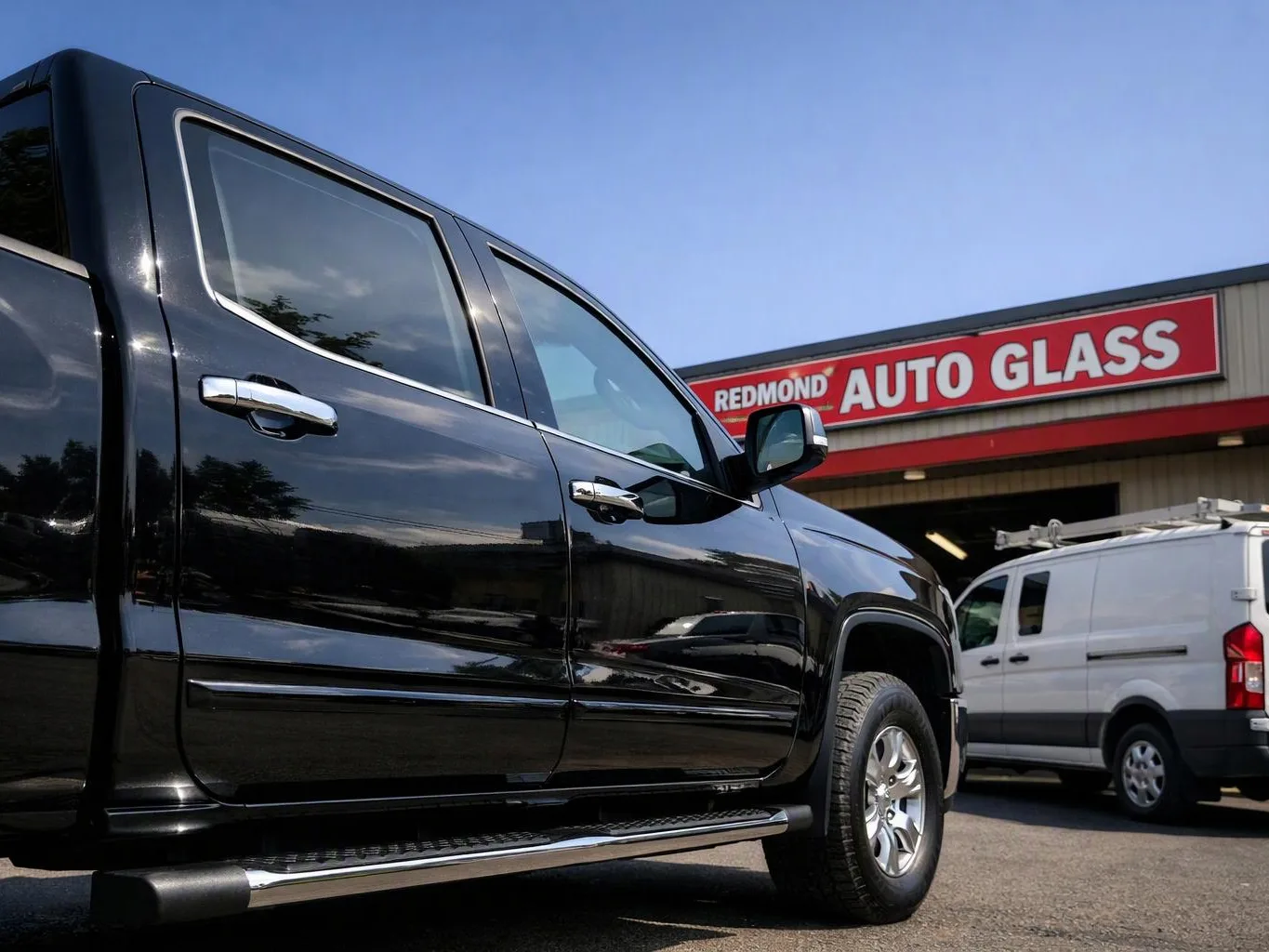A black pickup truck is parked outside an auto glass shop beside a white van
