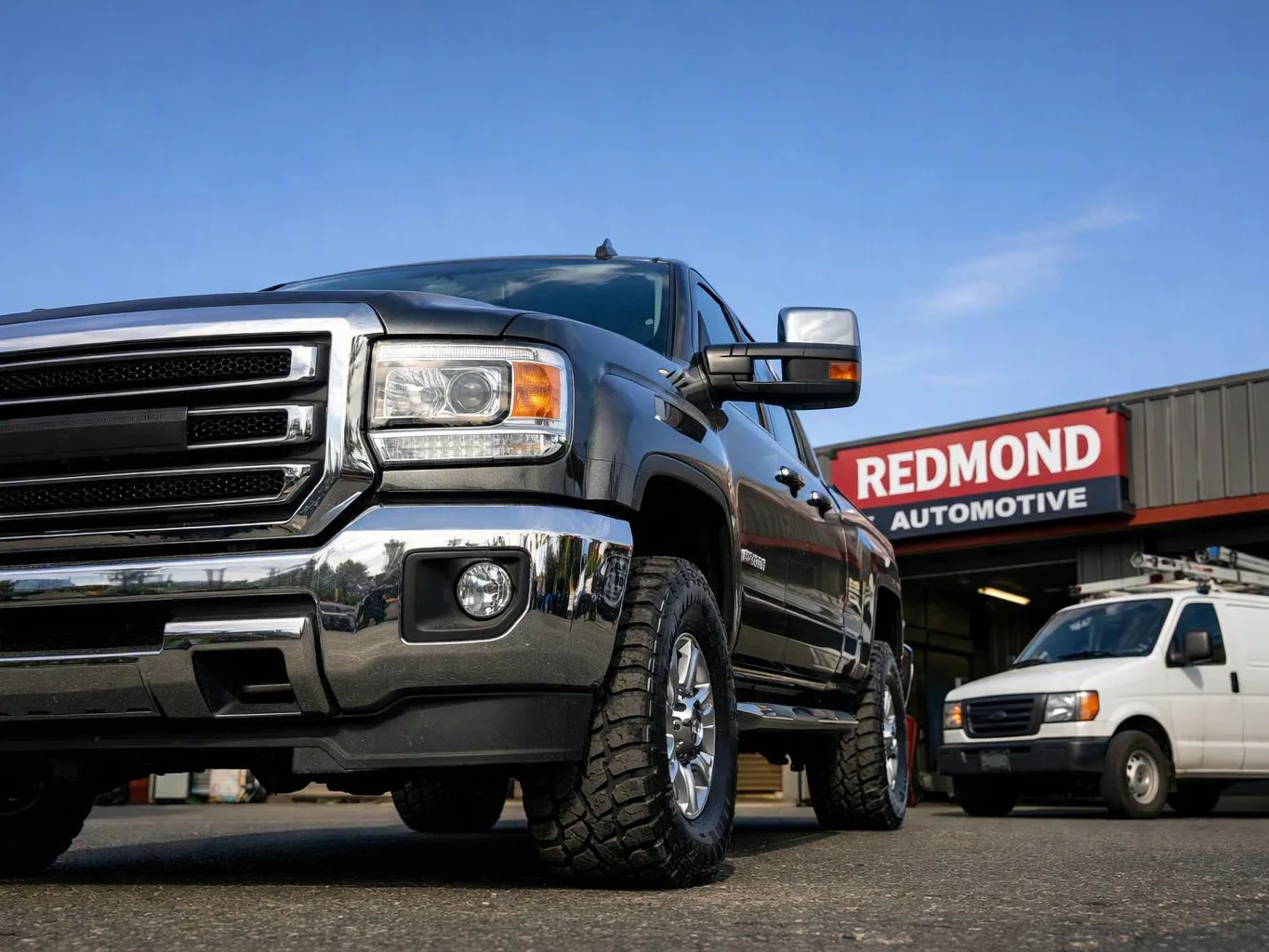 A black pickup truck is parked outside a garage beside a white work van under a clear blue sky