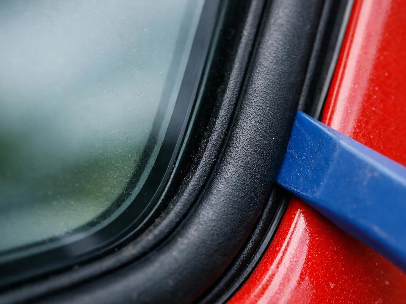 A blue pry tool lifts a black rubber seal beside a foggy car window and red body panel