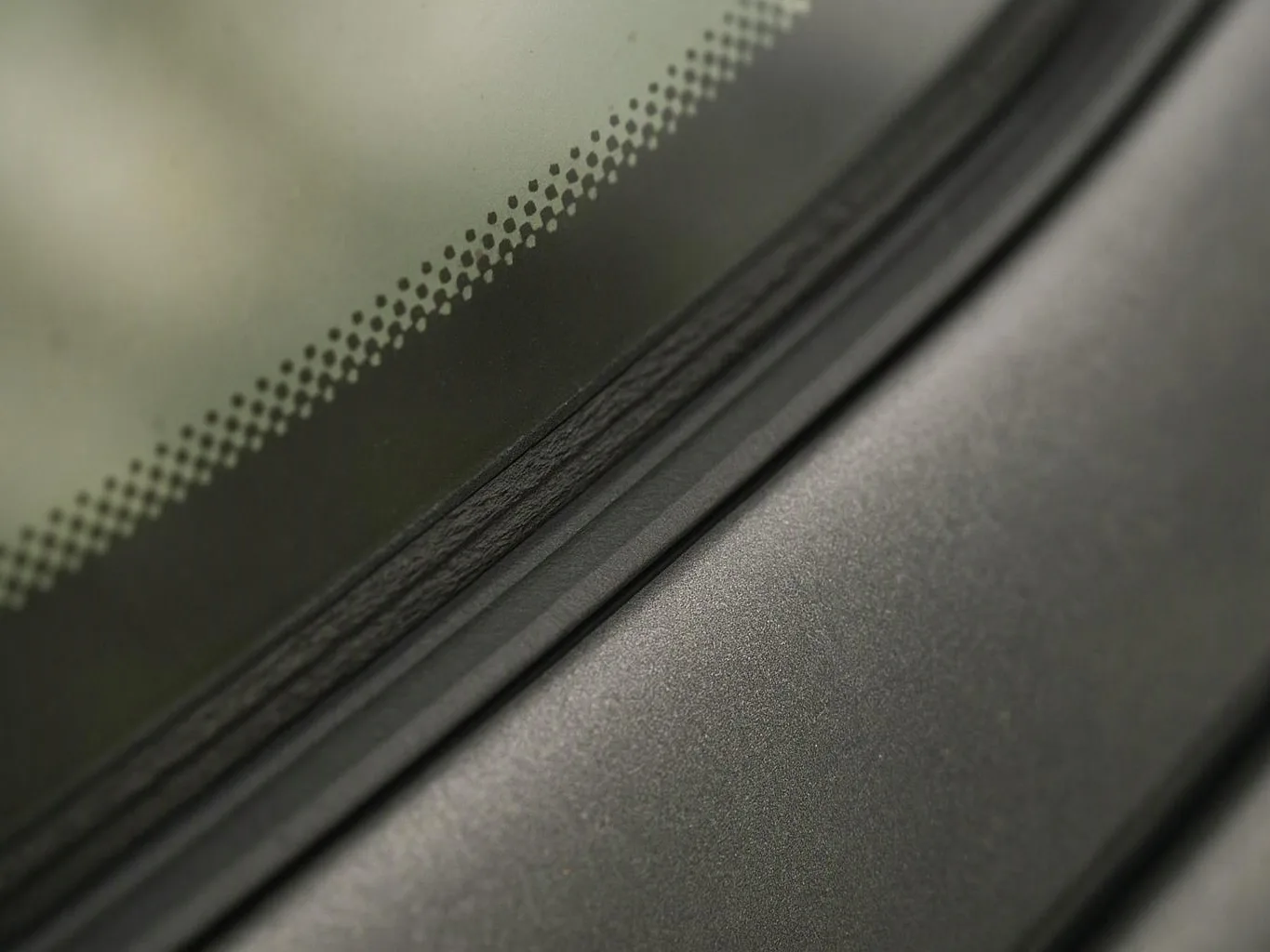 Close-up of a car windshield edge with dotted tint, black trim, and a gray hood surface