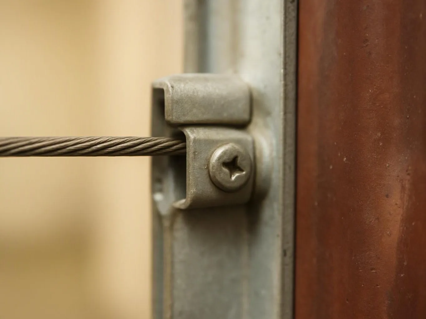 A steel cable passes through a metal clamp fastened to a brown wooden panel against a blurred background